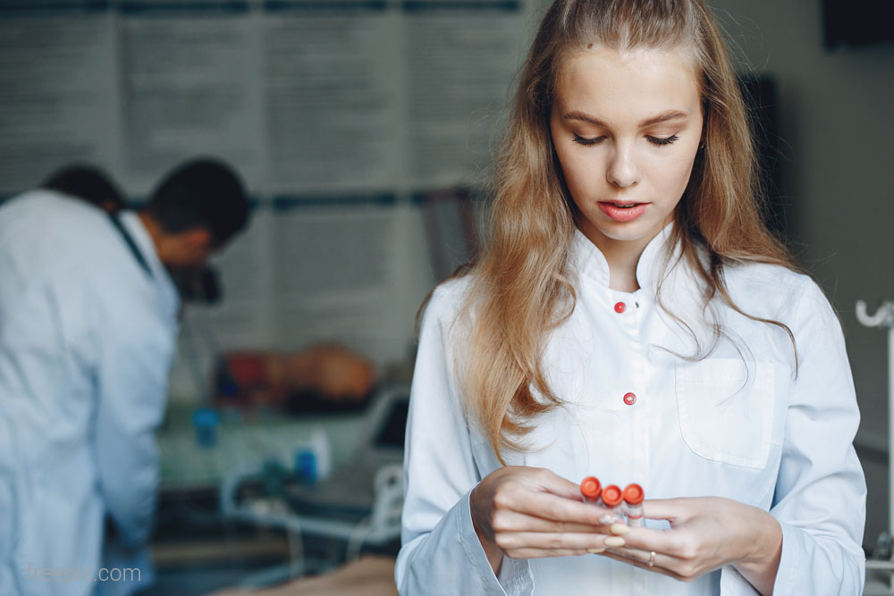 zdjęcie przedstawia pielęgniarkę, a w tle zdjęcia widoczni są pracownicy medyczni